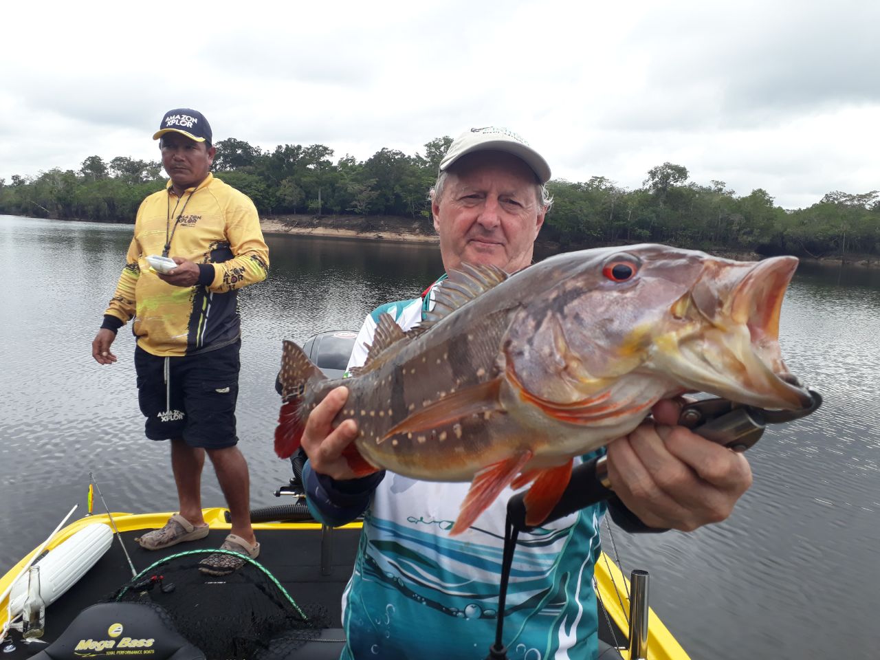 Fisherman in a cap showing a beautiful peacock bass he caught on Amazon Xplor wiht the fihing guifd in the back